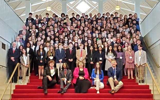 Group photo with the members of the 2024 Youth Legislative Assembly standing on a staircase lined with red carpet and gold hand rails